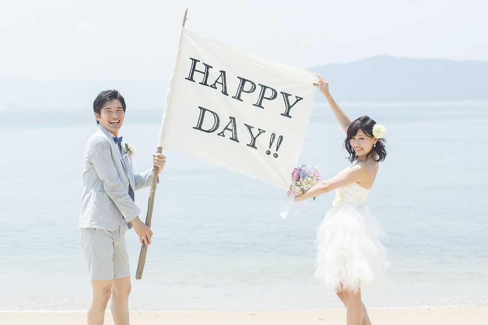 HAPPYDAYのフラッグを掲げて海辺で写真を撮る新郎新婦