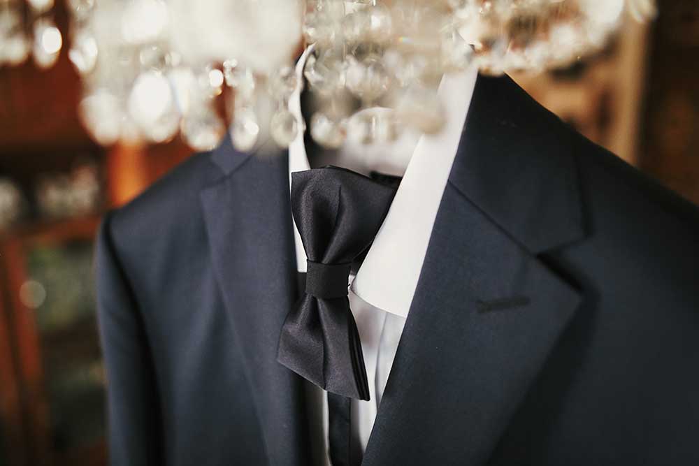 stylish black suit with white shirt and bow tie on hanger on luxury chandelier. morning preparations before wedding day. groom outfit.